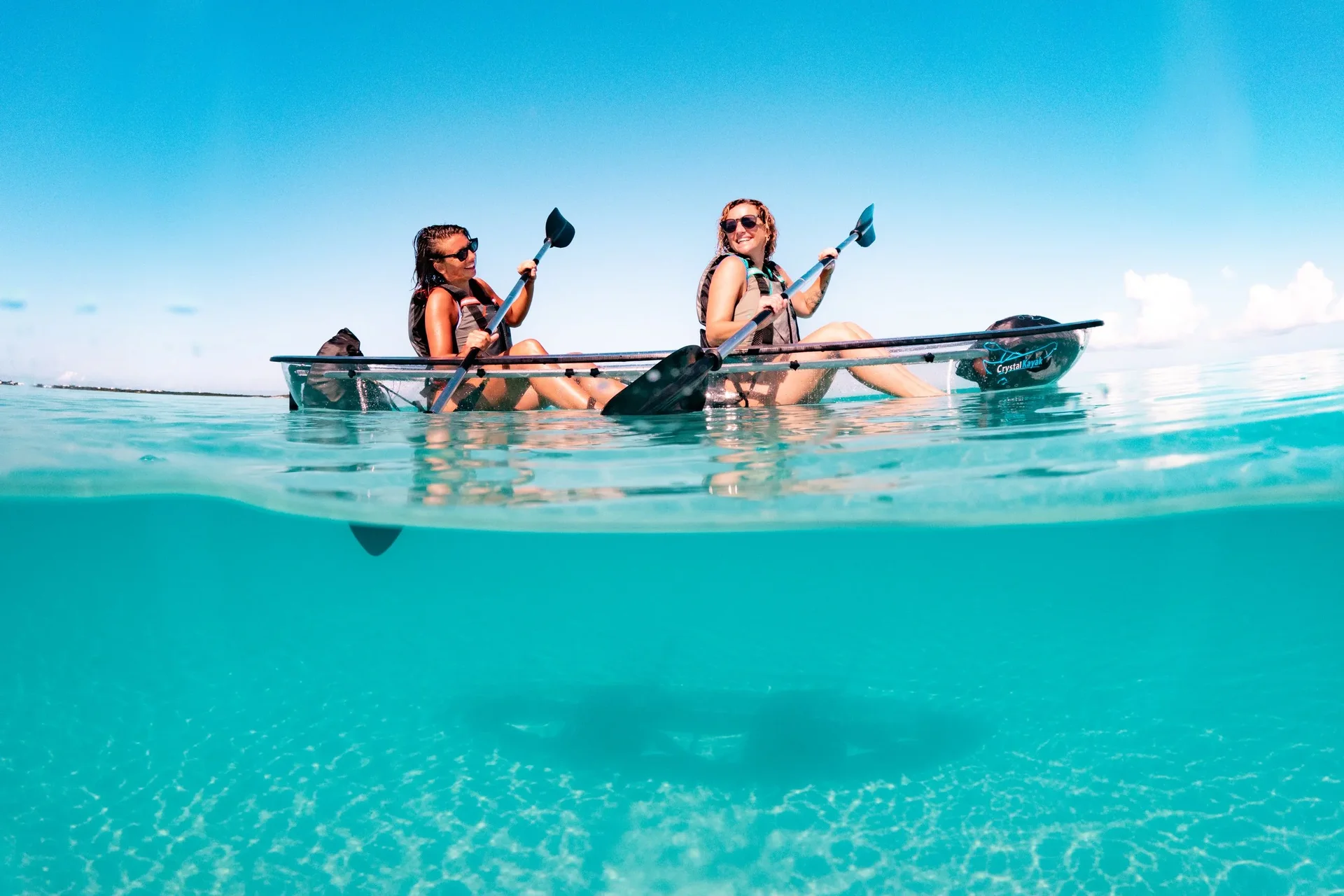 Two guests paddling a clear kayak over bright turquoise water